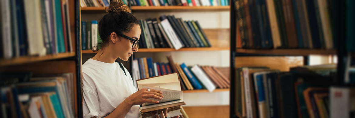 Woman standing in between bookstacks with an open book in hand