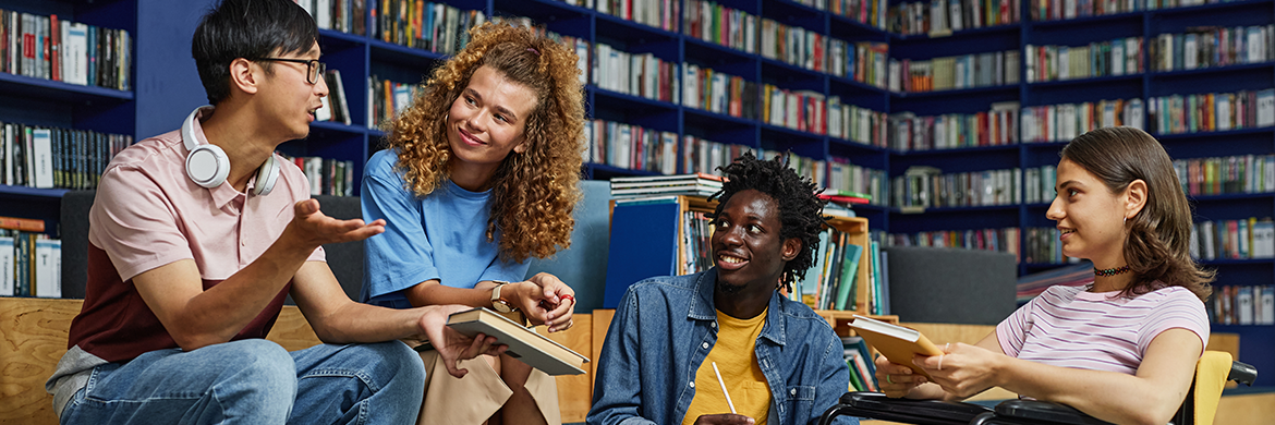 A group of four teens in discussion in a library setting.