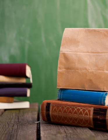 Two book stacks on a wooden table the stack of two books on the right has a brown lunch bag on top.  Green background