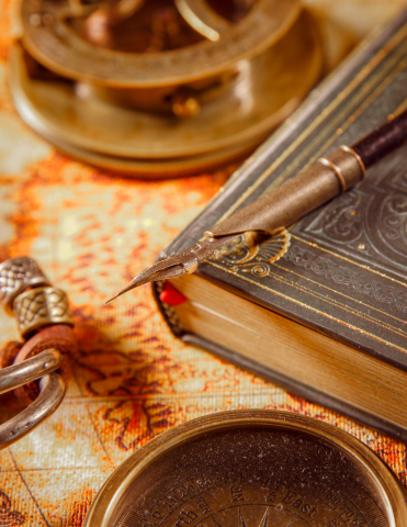 antique book with antique pen laying on top.  Book is resting on an old map with a compass and other old items surrounding