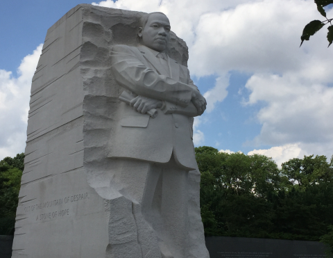 Martin Luther King statue with blue sky, white puffy clouds and a row of green trees in the background