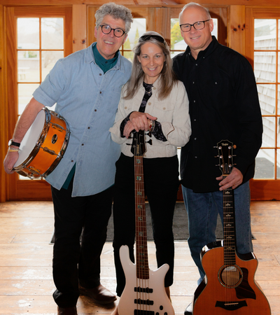 Three musicians holding instruments, a drum, a bass and a guitar in front of glass windows.