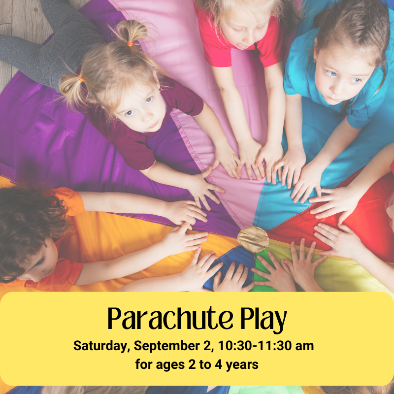 A group of seven kids, laying on their stomachs on top of a rainbow parachute in a circular formation.