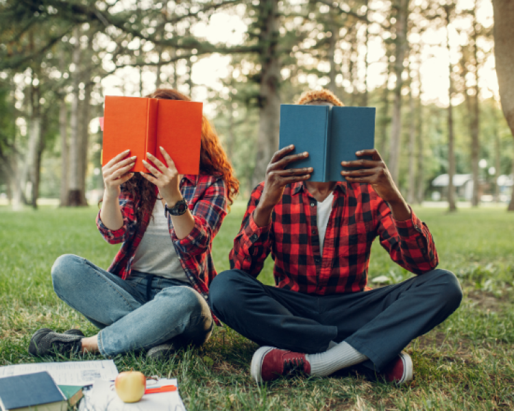 Two teens holding a book in front of their faces while sitting in the grass